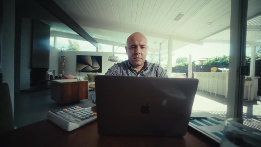 A man opens a laptop and looks at the screen while sitting at a desk in a modern home
