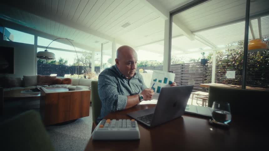 A man sits at a dining table in a modern sunlit room while working on a laptop he holds up a printed document featuring bar charts and uses a pen to point at specific data points