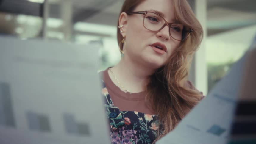 A young woman wearing glasses sits outdoors holding some documents with a computer in front of her