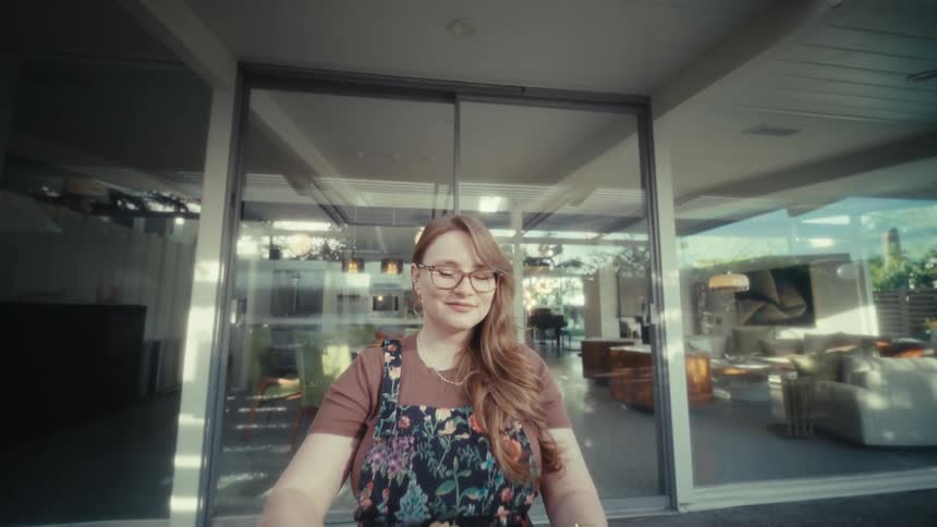 A woman with reddish hair and glasses smiles at the camera before opening a striped laptop on a wooden desk outside a modern glass house with a visible living room