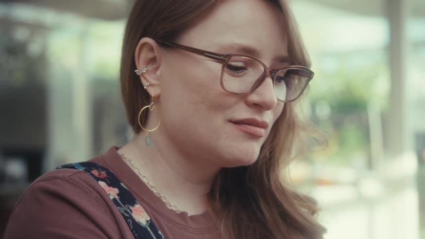 A woman with glasses and long reddishbrown hair is shown using a colorful laptop in an outdoor setting the video focuses on her face as she types eventually transitioning into a closeup that highlights her jewelry and a subtle smile