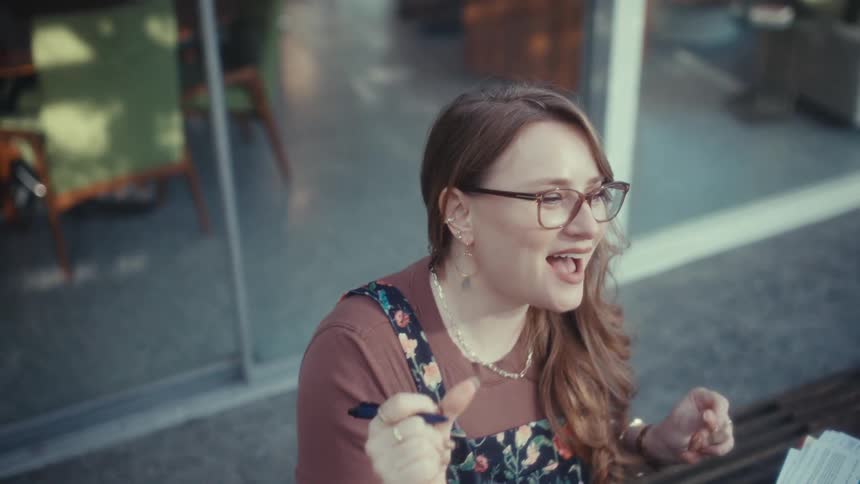 A woman wearing glasses and a floral patterned top sits in front of an open laptop while dancing and smiling during a video call at a wooden table in a bright modern setting