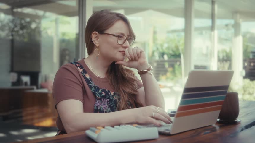 A woman with glasses and long hair sits at a wooden table on a bright patio typing on a laptop with a colorful sticker while pausing briefly to rest her chin on her hand in thought