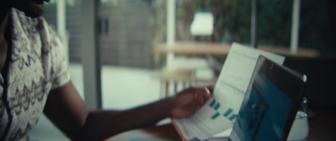 A man in a patterned shirt sits at a desk looking over printed charts and documents while working on a laptopthe camera transitions from a closeup of his focused expression to a view of his hands typing on the keyboard