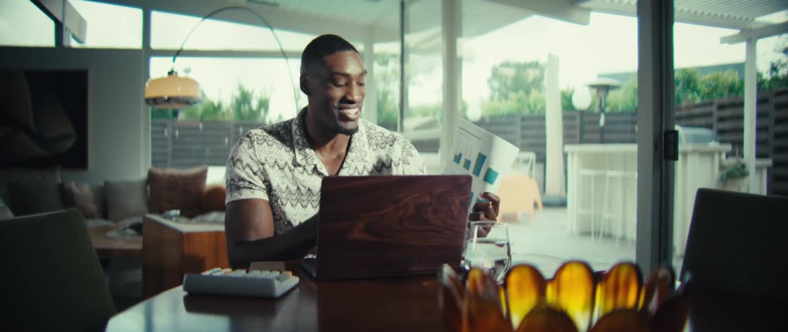 A man wearing a patterned short sleeve shirt sits at a wooden table and holds up a document with several colorful bar charts while speaking during a video call on a laptop in a bright modern home