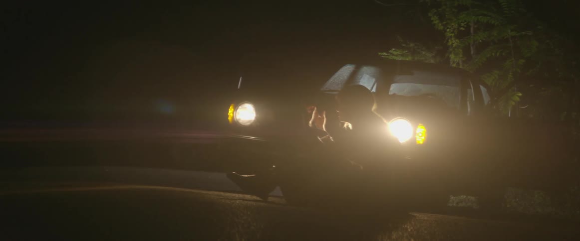 A woman sits in front of a car with its headlights on at night