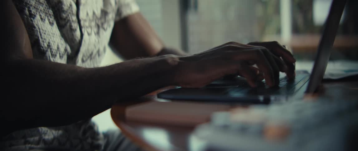 A closeup side profile of a persons hands typing on a laptop keyboardthe individual is seated at a wooden table in a dimly lit indoor setting with a shallow depth of field focusing on the movement of their fingers