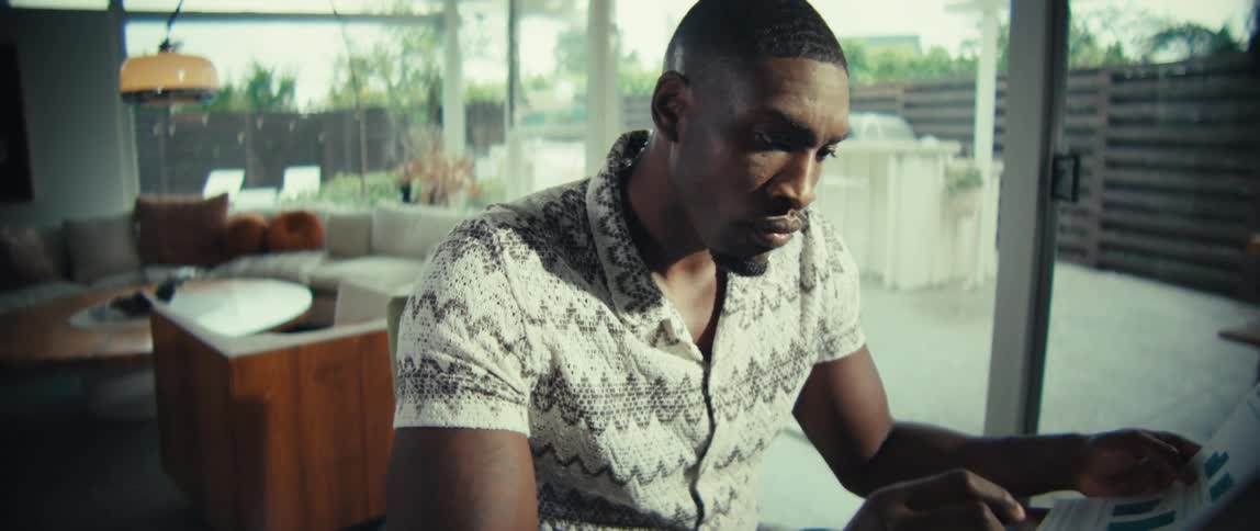 A black man is sitting at a desk in a modern living room working on a laptop in a sunlit room