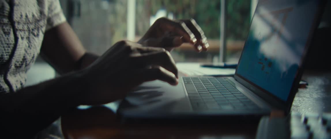 A man holds a paper with bar charts while looking at a laptop screen before setting the document down to type on the keyboard at a wooden desk near a bright window