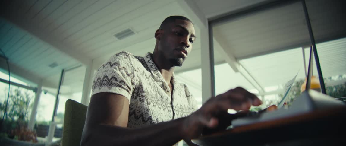 A man with dark skin wearing a patterned shirt sits at a desk and types on a white calculator in a bright room