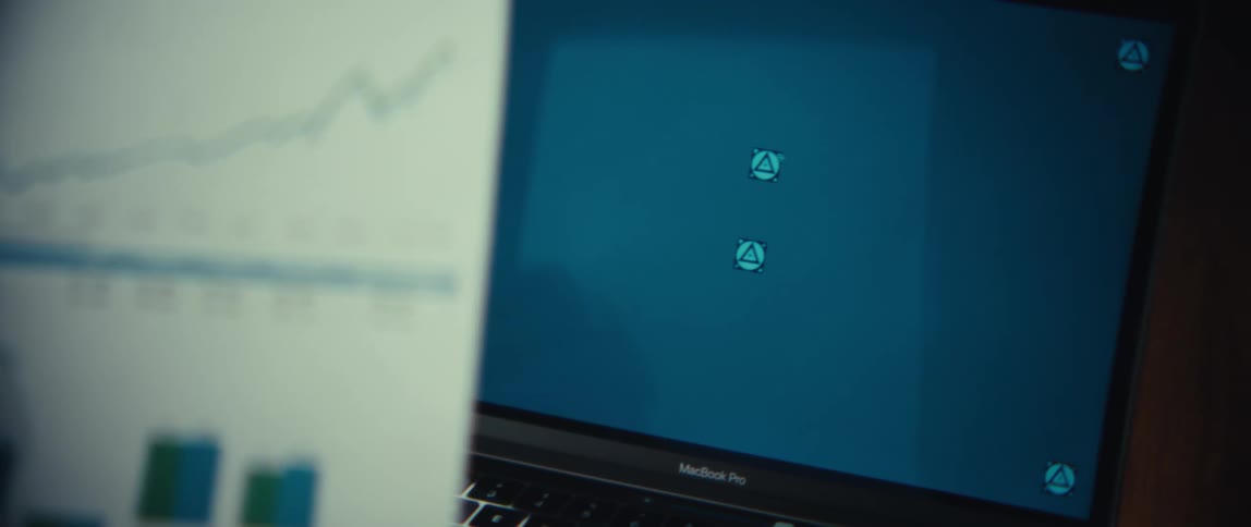 A person holds a paper report showing a portfolio growth chart and financial data tables while sitting in front of a laptop screen displaying blue icons in a dimly lit office environment