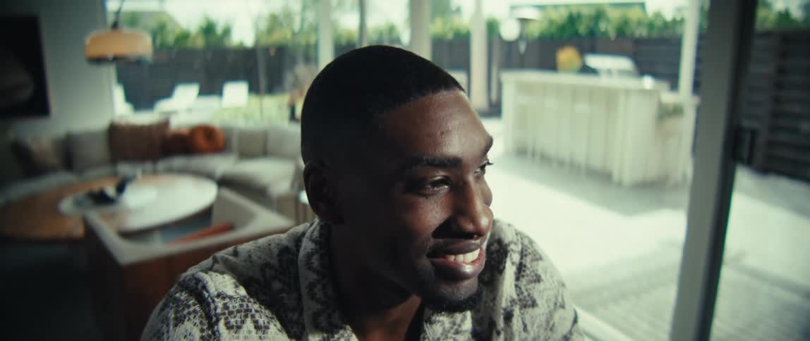 A hapy black man sits at a table near a large window looking thoughtfully while working on a laptop with charts and documents spread out beside him