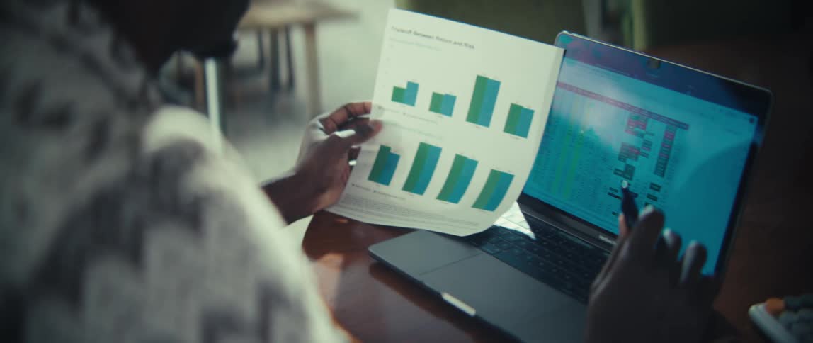 A man in a patterned shirt analyzes a bar chart titled tradeoff between return and risk while holding a pen near his laptop the computer screen displays a colorful data grid or heatmap that he appears to be comparing with the printed document
