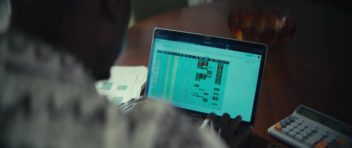 A close view of a man working on a laptop with a spreadsheet on the screen at a wooden table in a bright modern home