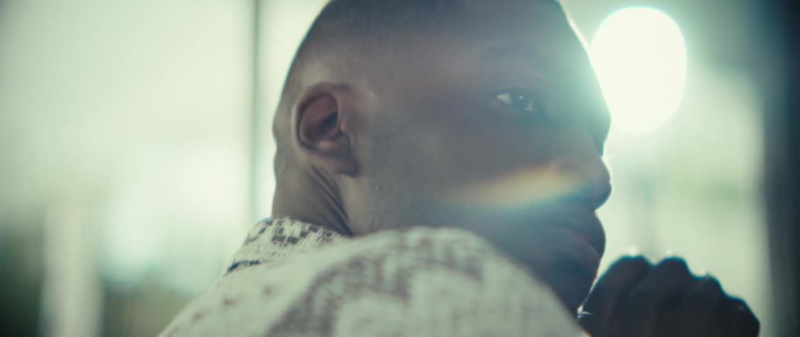 A young black man sits indoors pausing for a moment with his hand on his jaw in deep thought as sunlight streams through the glass windows