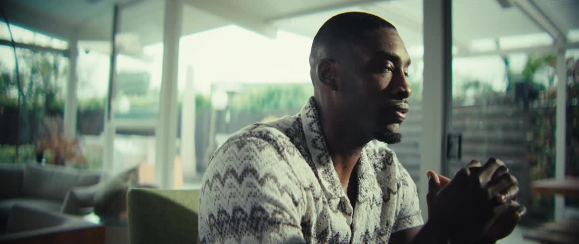 A man wearing a patterned short sleeve shirt sits at a dining table and types on a laptop before looking up and toward the side in a bright modern home with glass walls