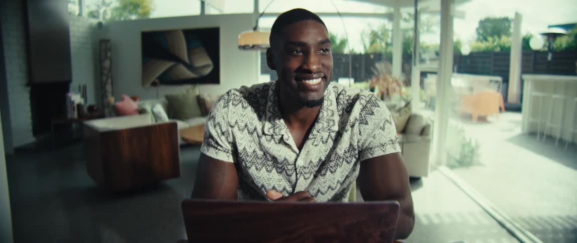 A man in a patterned shirt works on a laptop at a table featuring a calculator and chartshe begins with a focused expression before looking up with a wide smile and returning to his typing