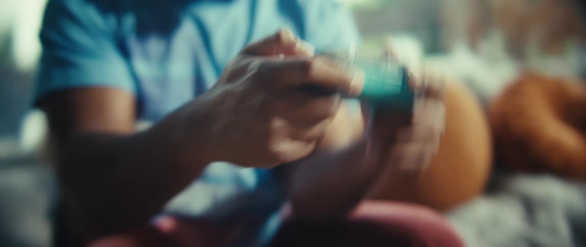 A black man sits on a couch in a sunlit living room using a game controller excitedly