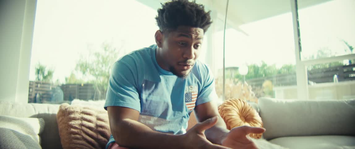 A black man is sitting on a couch in a modern living room waving and talking towards a tablet on a table near him