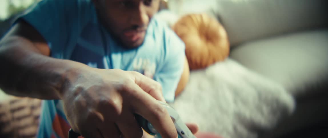 A man wearing a blue shirt plays a video game with a controller while sitting on a sofa in a bright living room in a close view