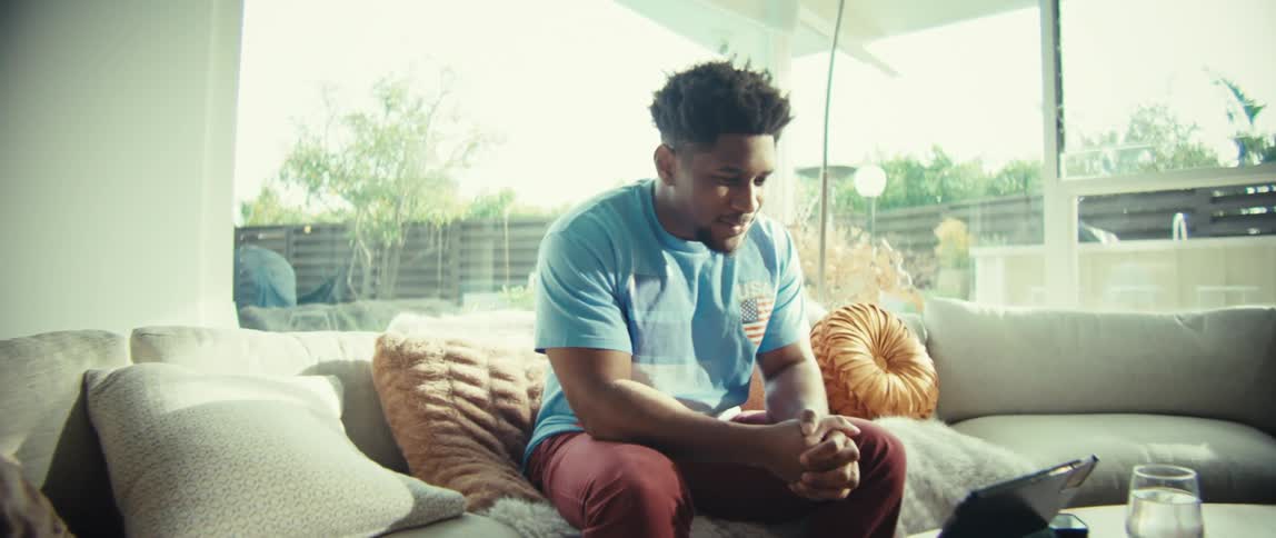 A man wearing a blue shirt sits on a sofa and looks at a tablet on a table while speaking and moving his hands in a bright living room