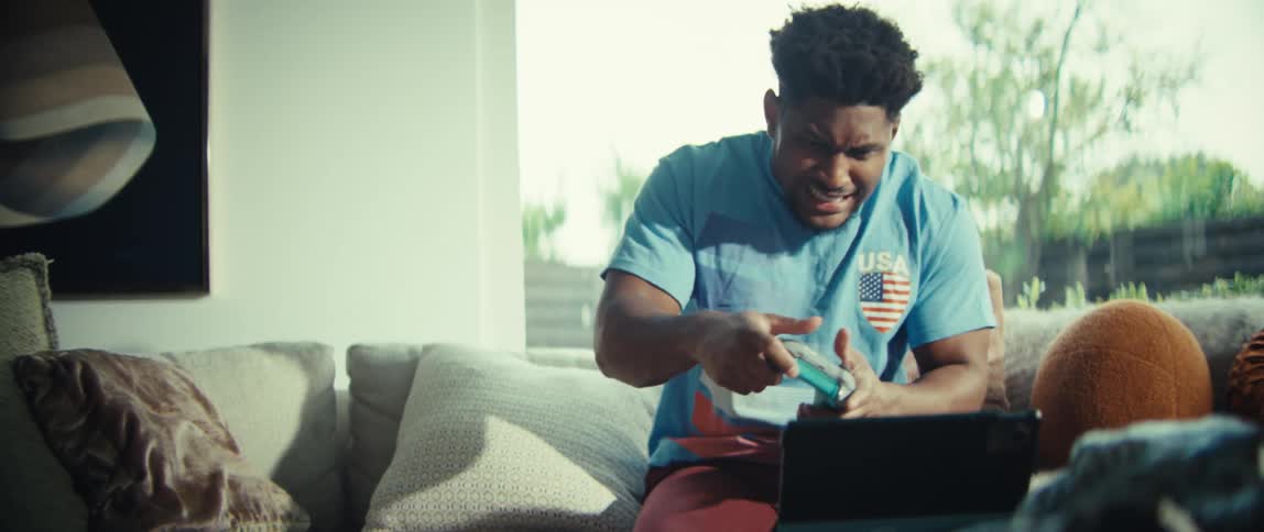 A man in a usa tshirt sits on a couch while playing a video game on a tablethe reacts with intense excitement shouting and pumping his fist as he celebrates a victory