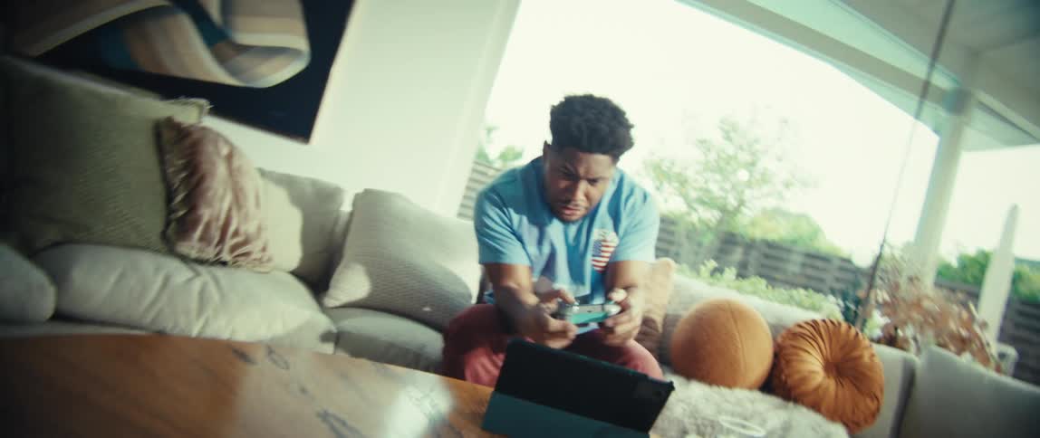 A young man sits on a couch playing a video game excitedly at midday