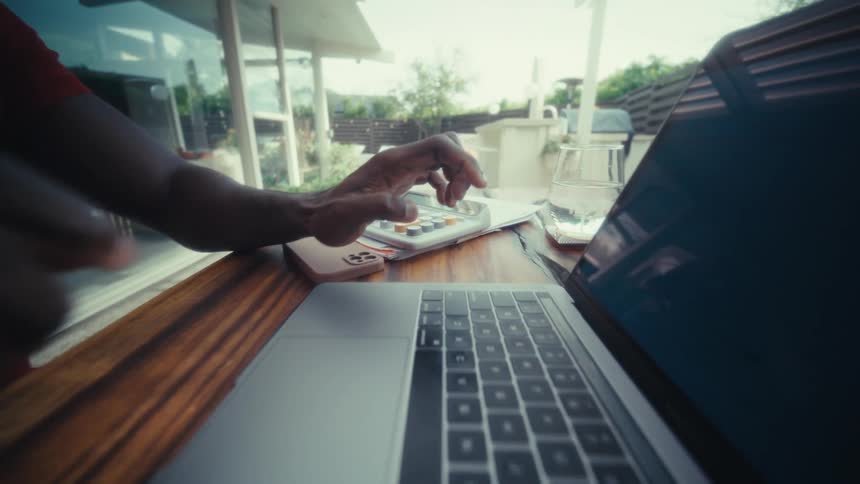 A close view shows the hands of a man as he types on a laptop keyboard at a wooden table in a bright outdoor patio area