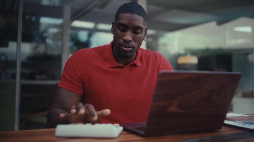 A man seated at a table outside uses a calculator and a laptop