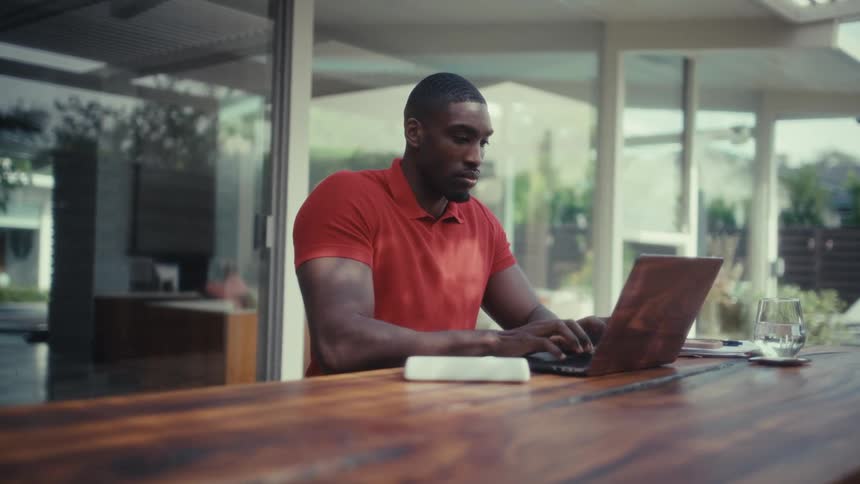 A man wearing a red polo shirt types on a laptop at a long wooden table in a bright modern home with glass doors leading to an outdoor patio