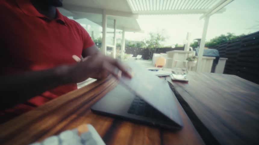 A man wearing a red polo shirt types on a laptop at a wooden table before closing the screen and looking toward the side with a smile in a bright outdoor patio area