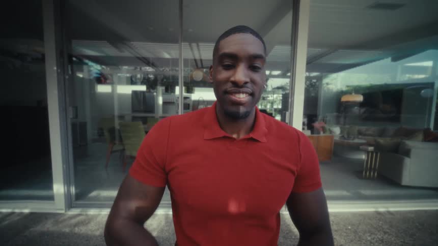 A man wearing a red polo shirt opens the lid of a laptop and smiles while looking directly at the camera in a bright modern home