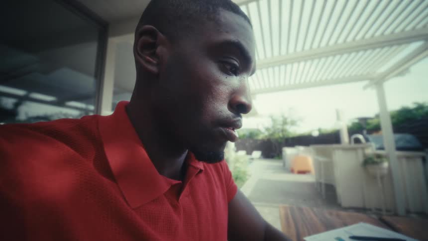 A man in a red polo shirt sits at a wooden outdoor table and opens his laptop to begin workingthe camera moves into a closeup of his face as he looks at the screen with a focused expression