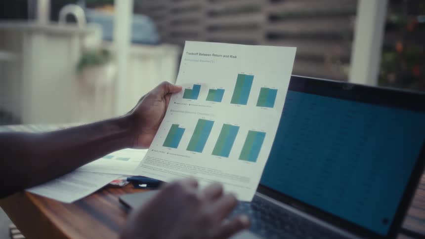 Man compares computer data and a printout while working at a desk during the daytime