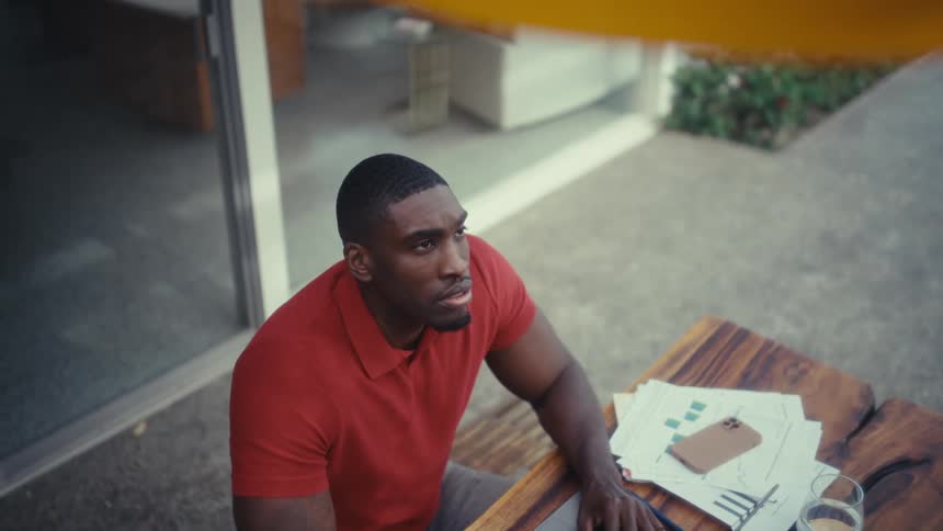 The video shows a man in a red polo shirt sitting at an outdoor wooden table while working on a laptop after typing for a moment he pauses looks up toward the sky and begins to smile