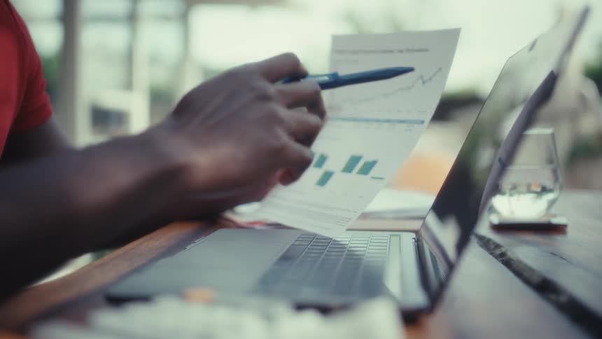 A man points a blue pen at a financial growth chart while sitting at a wooden table with a laptop