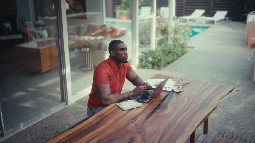 The man is typing on a laptop at a large table in his yard