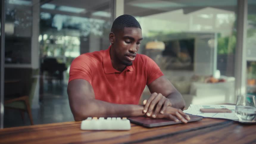 A man wearing a red polo shirt speaks while looking at a laptop and using a calculator while sitting at a large wooden table in a bright modern home