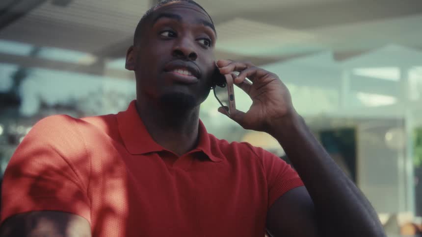 A man wearing an orange polo shirt sits at a desk while talking on a smartphone and looking at a laptop he starts the conversation with a neutral expression but quickly breaks into a wide joyful smile as he continues to speak