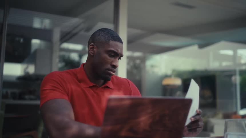 Man seated at a table uses a laptop while holding a document beside it