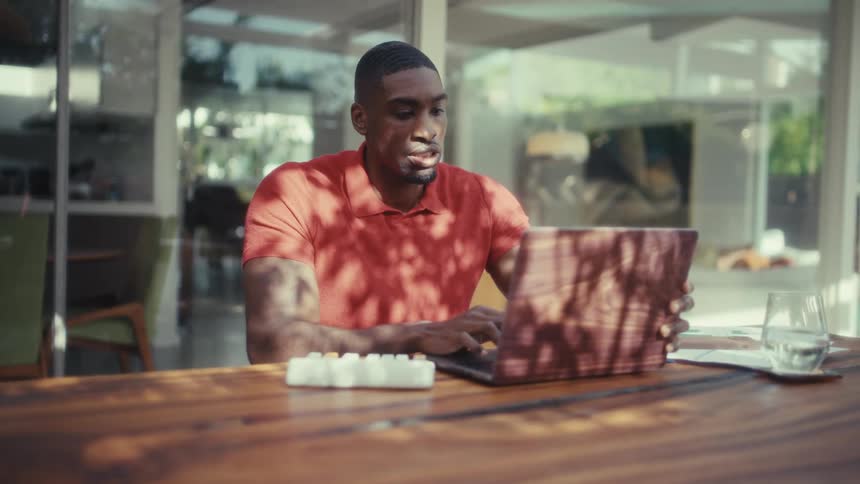 A young black man sits in an outdoor dining area at midday typing on a laptop before suddenly closing the lid in sadness