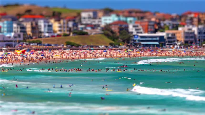 Crowded beach with swimmers in the sea and buildings in the background under a blue sky