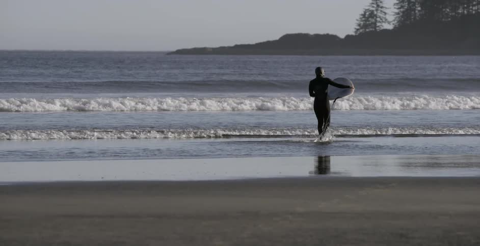 Man in wet suit holding surfboard walks out into ocean water