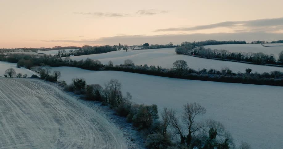 Snow covers fallow fields enclosed by trees in the rural countryside under a red sky