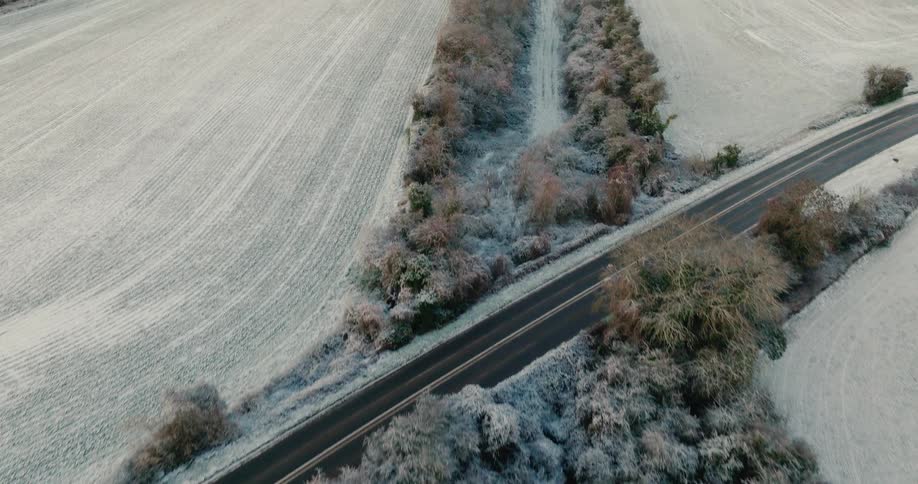 An aerial view shows snow covered fields and frost tipped trees surrounding a small road with cars passing by under a pale winter sky at dusk