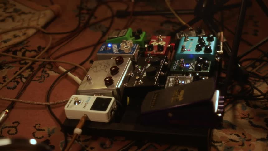 A cluster of colorful guitar effects pedals sits on a pedalboard atop a patterned rug with cables loosely coiled around them