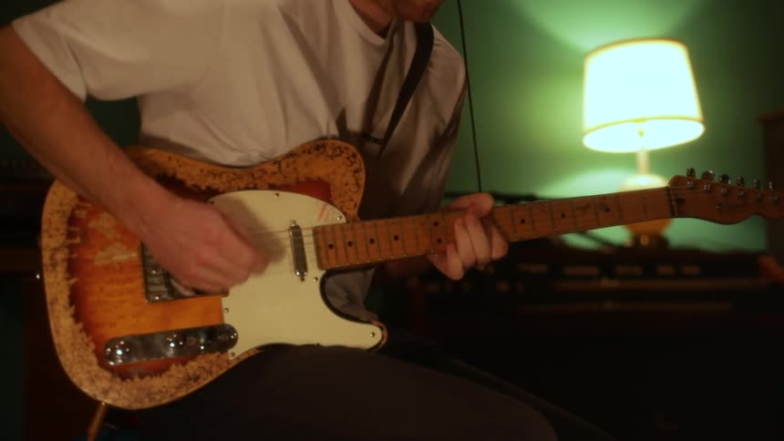 A man wearing a white tshirt sits in a dimly lit room with a glowing lamp and plays a sunburst electric guitar with visible wear on the body