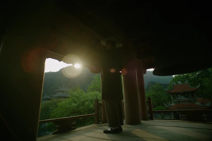 A monk stands inside a temple at sunset wearing a hat with the surroundings covered in green vegetation