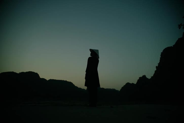 A monk wearing a hat stands on mountainous terrain at night