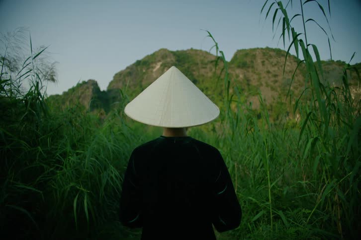 A man wearing a traditional conical hat and black clothing stands with his back to the camera looking toward large green mountains and rocky cliffs surrounded by dense tall grass and lush tropical vegetation under a clear sky